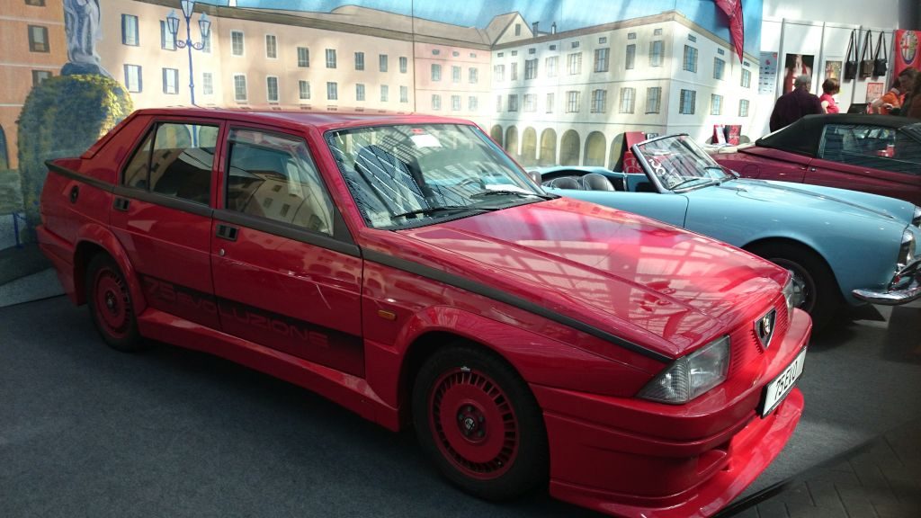 Ein roter Alfa Romeo wird im Innenbereich vor einer gemalten Gebäudekulisse neben einem hellblauen Cabrio ausgestellt und spiegelt so die Atmosphäre der Techno Classica Essen 2015 wider.