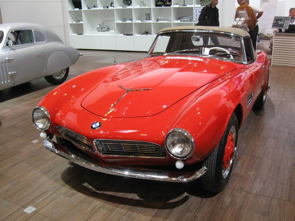 Ein rotes Vintage-Cabriolet von BMW wird in einem Ausstellungsraum ausgestellt, im Hintergrund sind andere Fahrzeuge und Autoteile zu sehen, was an die exquisiten Ausstellungen der Techno Classica Essen 2015 erinnert.