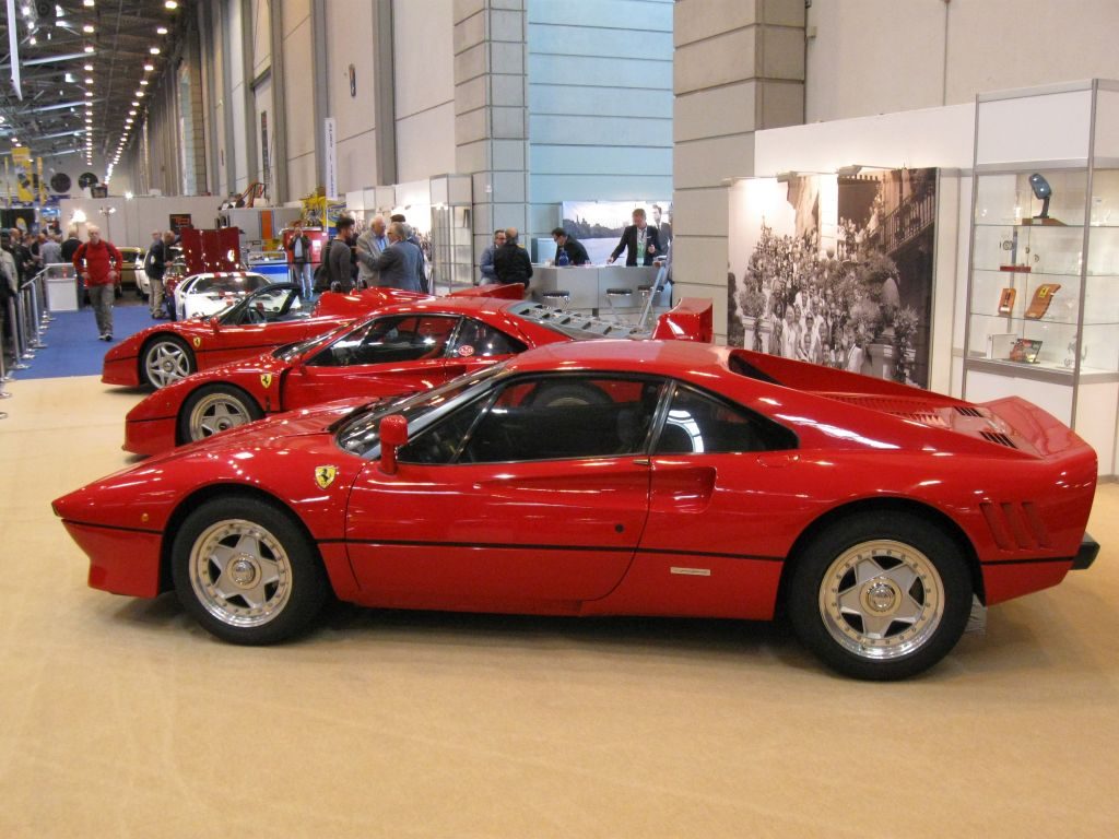 Drei rote Ferrari-Sportwagen werden im Innenbereich der Techno Classica Essen 2015 ausgestellt. Im Hintergrund einer Ausstellungshalle laufen Menschen herum und weitere Stände sind zu sehen.