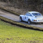 Ein weißer klassischer TANDLER Porsche 911 RSR fährt auf einer schmalen Landstraße, die von Gras und blattlosen Bäumen gesäumt ist.