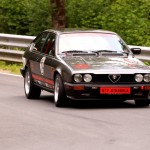 Ein schwarzer Alfa Romeo GTV Stradale, der mit einer Metallleitplanke an der Seite auf einer Waldstraße fährt, weckt den Nervenkitzel des Wolsfelder Bergrennens.