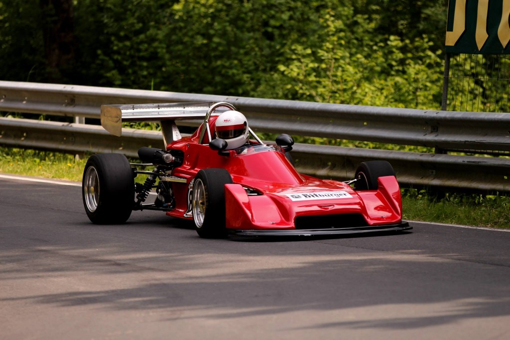 Eine Person in einem roten Formel-Rennwagen mit Helm und Rennmontur fährt auf der Strecke des Wolsfelder Bergrennens, die von einer Metallleitplanke und grünem Laub begrenzt wird.