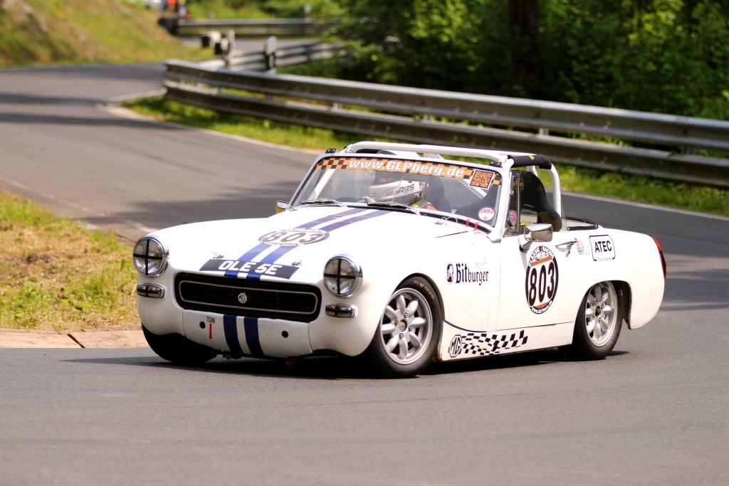 Ein weißer, klassischer Rennwagen mit der Startnummer 803 rast durch die kurvenreichen Straßen des Wolsfelder Bergrennens, gesäumt von Metallleitplanken und Bäumen. Auf der Motorhaube und den Seiten des Wagens sind Aufkleber und Sponsoren zu sehen, die seinem eleganten Design Flair verleihen.