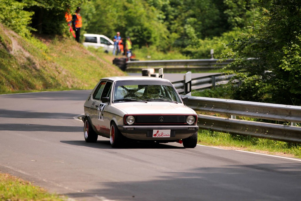 Ein weißer Oldtimer mit der Nummer 52 fährt auf einer kurvenreichen Straße umgeben von Grün, die an das Wolsfelder Bergrennen erinnert, im Hintergrund sind Zuschauer und Offizielle zu sehen.