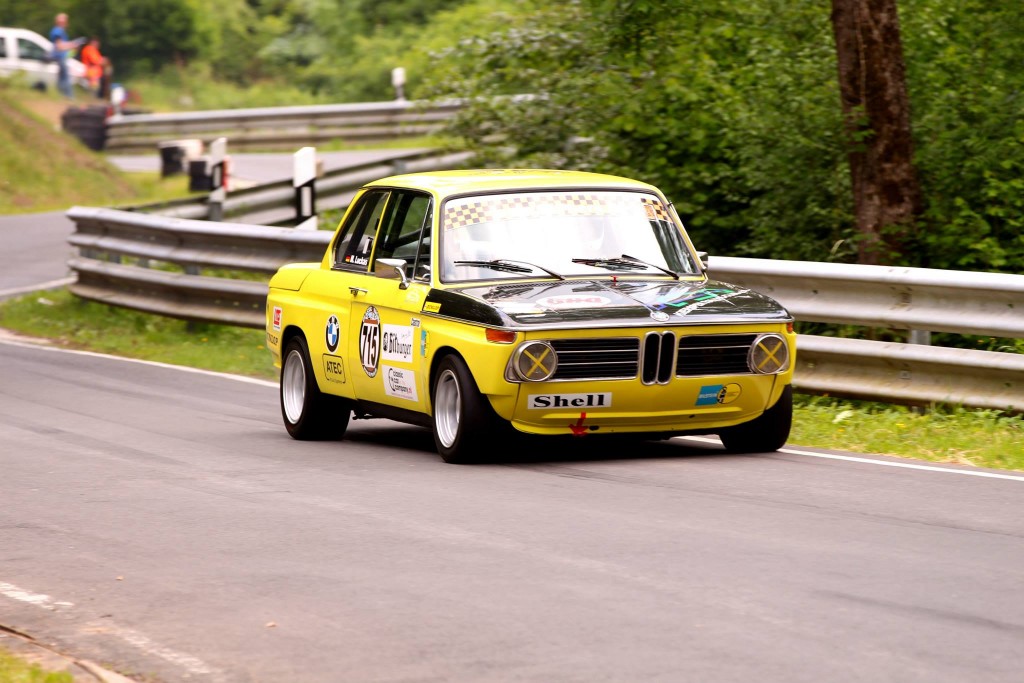 Ein gelb-schwarzer klassischer BMW-Rennwagen, geschmückt mit verschiedenen Sponsorenaufklebern, darunter auch Shell, rast durch die kurvigen Straßen des Wolsfelder Bergrennens mit Leitplanken und üppigem Grün im Hintergrund.