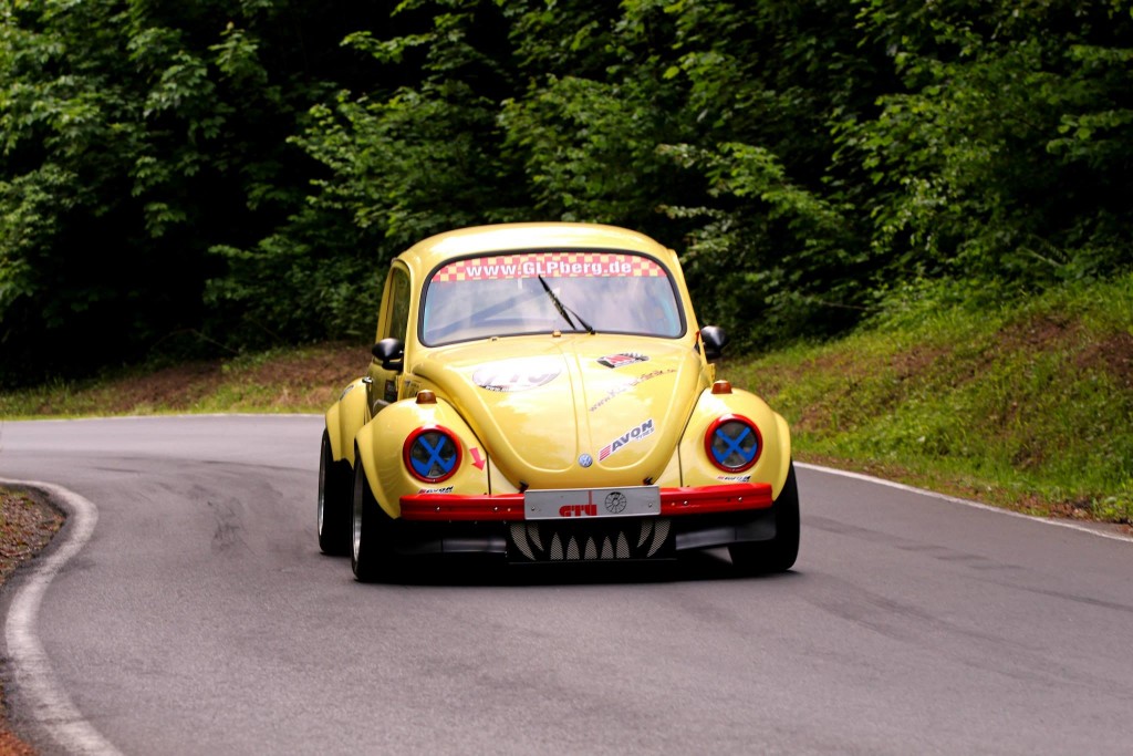 Ein gelber, modifizierter VW Käfer mit Aufklebern fährt auf einer kurvigen, von Bäumen gesäumten Straße, die an das Wolsfelder Bergrennen erinnert.