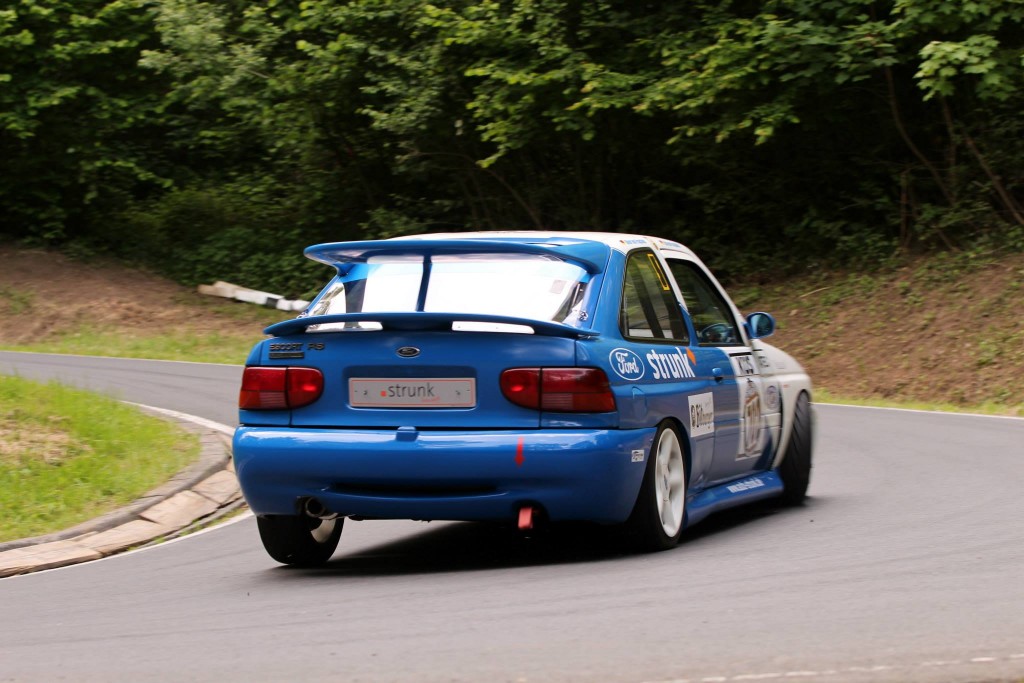 Ein blau-weißes Rennauto mit verschiedenen Sponsorenlogos fährt beim Wolsfelder Bergrennen auf einer kurvigen, asphaltierten Straße, die von grünen Bäumen umgeben ist. Das Auto hat einen großen Heckspoiler und ist von hinten zu sehen.