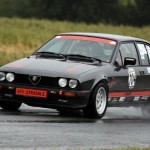 Ein schwarzer Alfa Romeo GTV Stradale mit Rennaufklebern fährt beim Hauenstein Bergrennen 2015 auf einer nassen Strecke und spritzt dabei wie eine Wasserspritzer hinter sich her.