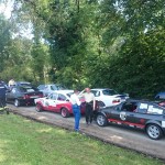 Eine Reihe von Oldtimern und klassischen Autos, die entlang einer von Bäumen gesäumten Straße geparkt sind, während Menschen an einem sonnigen Tag in der Nähe der Fahrzeuge stehen und gehen, erinnert an den Charme von Veranstaltungen wie dem 48. Internationalen Osnabrücker ADAC Bergrennen.