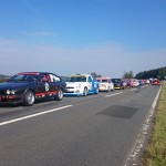 Eine Reihe bunter Autos, die an das Bergrennen Unterfranken 2015 erinnern, parkt unter einem klaren blauen Himmel am Rand einer Landstraße, im Hintergrund sind Felder und Bäume zu sehen.