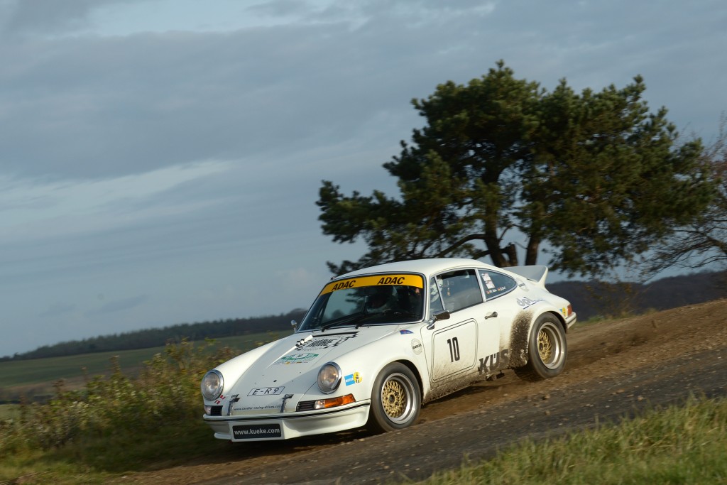 Ein weißes Rennauto mit der Nummer 10 auf der Seite, das an der Rallye Köln-Ahrweiler teilnimmt, fährt auf einer Landstraße mit einem Baum und einem Feld im Hintergrund.