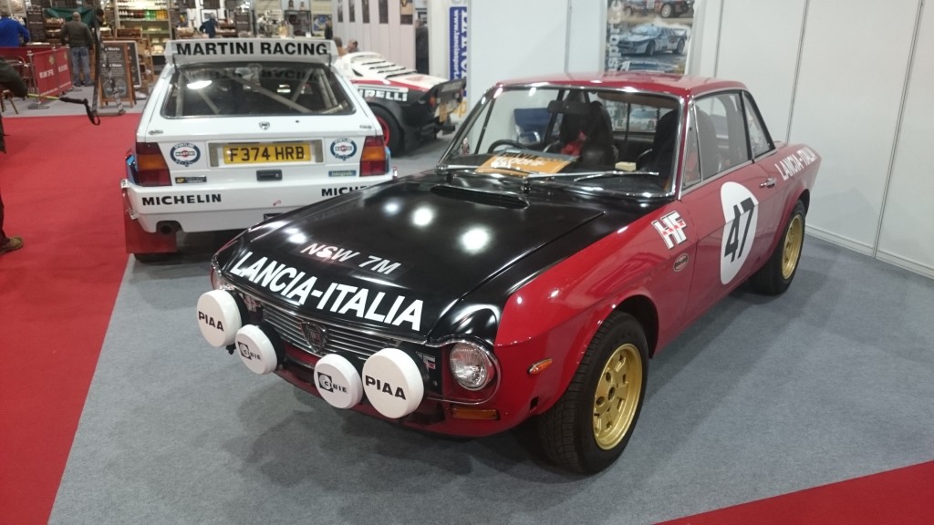 Ein klassisches rot-schwarzes Lancia-Rallyeauto mit der weißen Nummer 41 wird bei Race Retro 2016 neben einem Martini-Rennauto auf der Indoor-Autoshow ausgestellt.