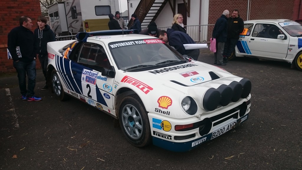 Ein weißes Rallyeauto mit der Nummer 2 und mehreren Sponsorenaufklebern, darunter Shell und Pirelli, ist bei einer Veranstaltung im Freien während des Race Retro 2016 geparkt. Mehrere Personen haben sich um das Auto versammelt.