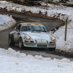 Ein klassisches Rallyeauto der Rallye 70 Kempenich fährt im Winter auf einer schneebedeckten, nassen Straße in einem Waldgebiet.