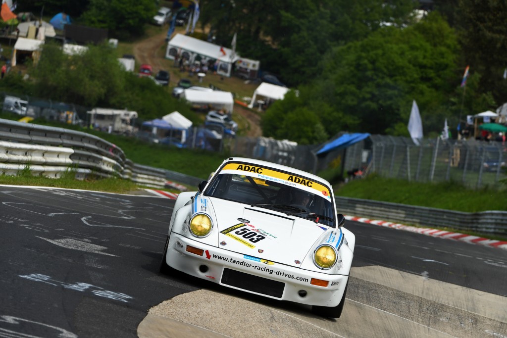 Ein weißer Rennwagen mit der Nummer 506 fährt auf einer kurvenreichen Rennstrecke mit grünen Hügeln und Zuschauerzelten im Hintergrund. Auf dem Auto sind Werbebanner angebracht, die an die 24 H Classic mit dem TANDLER Porsche 911 RSR erinnern.