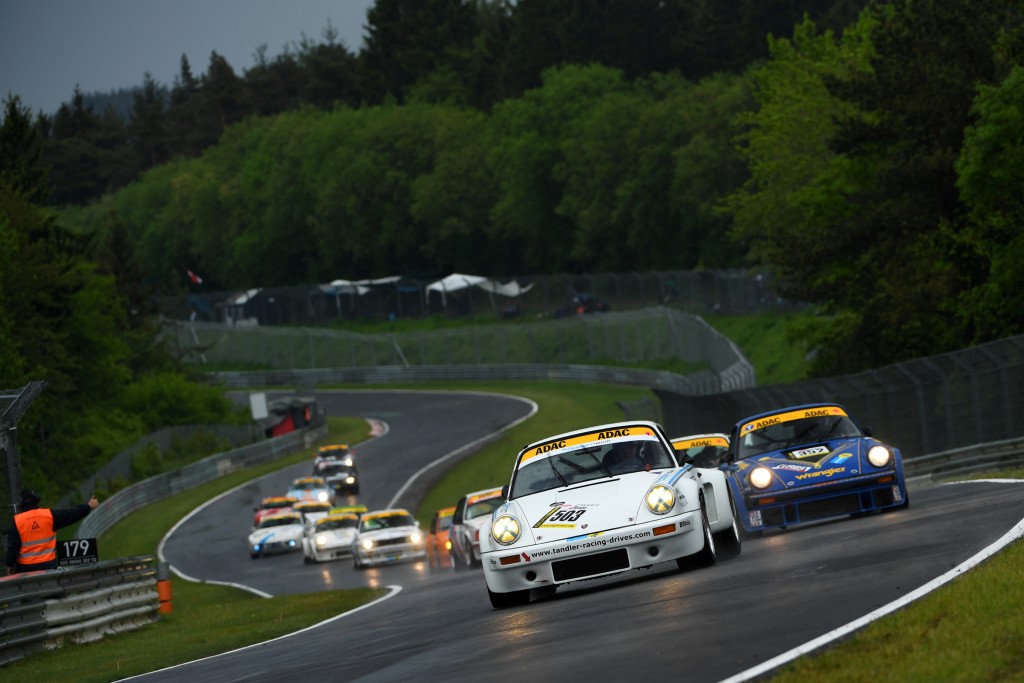 Eine Gruppe von Rennwagen, darunter der berühmte 24 H Classic mit dem TANDLER Porsche 911 RSR, navigiert eine kurvenreiche Strecke, die von üppigem Grün und Zuschauerzelten im Hintergrund umgeben ist. Das weiße Auto führt die Truppe durch dieses malerische Hochgeschwindigkeitsrennen an.