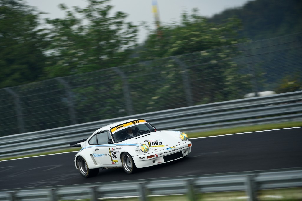 Ein weißer Rennwagen mit der seitlichen Startnummer 503 fährt im Rahmen der 24 H Classic mit dem TANDLER Porsche 911 RSR auf einer Rennstrecke, eingerahmt von Bäumen und Metallbarrieren im Hintergrund.