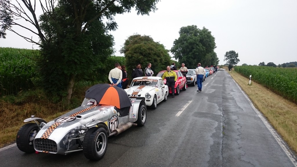 Eine Gruppe historischer Sportwagen mit Fahrern und Passagieren steht an einem bewölkten Tag auf einer Landstraße neben Feldern aufgereiht und erinnert an den Weserbergpreis 2016. Ein Auto im Vordergrund hat einen Regenschirm über der Fahrerseite.