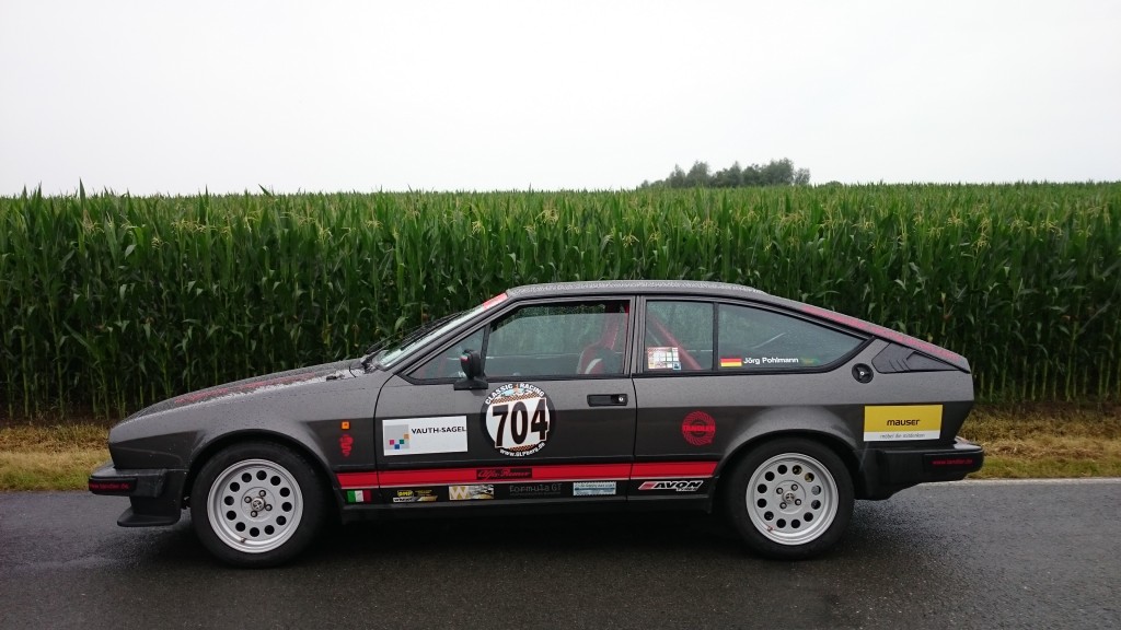 Seitenansicht eines grauen Alfa Romeo GTV6 mit Rennlackierung und der Nummer 704 auf der Tür, geparkt auf einer Straße mit einem Maisfeld im Hintergrund, aufgenommen während des Weserbergpreises 2016.