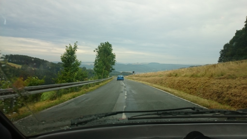 Ein Blick aus dem Inneren eines Autos auf einer schmalen Landstraße, vorn ein blaues Auto, umgeben von Bäumen und Feldern unter einem bewölkten Himmel – die Kulisse erinnert an die malerischen Routen beim Weserbergpreis 2016.