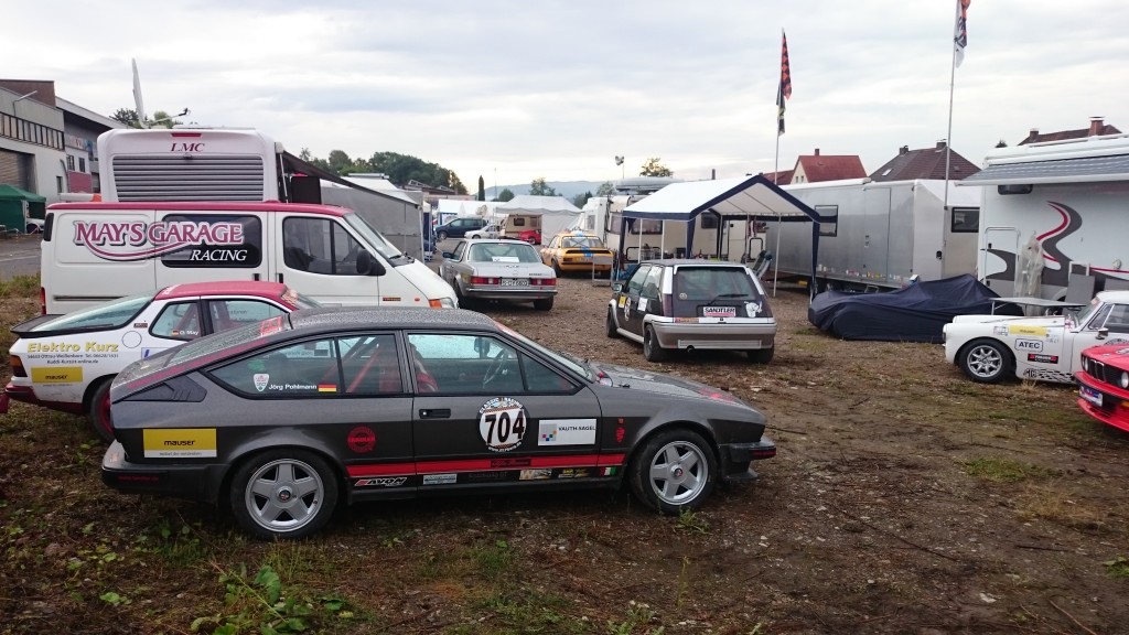 Auf einem Parkplatz steht ein Rennwagen mit der Aufschrift „704“ zusammen mit weiteren Fahrzeugen und Anhängern, darunter ein Lieferwagen mit der Aufschrift „May’s Garage Racing“. Im Hintergrund sind mehrere Zelte und ein wolkiger Himmel zu sehen, die an die Atmosphäre des Weserbergpreises 2016 erinnern.