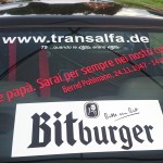 Heckscheibe des Autos mit der Website-URL „www.transalfa.de“, einer Gedenkbotschaft in italienischer und deutscher Sprache für „Bernd Pohlmann“ (24.11.1947 – 24.07.2015), einem „Osnabrücker Bergrennen 2016“-Aufkleber und einem „Bitburger“-Sticker.