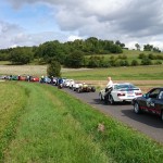 Eine Reihe geparkter Rennautos auf einer Landstraße mit grünen Feldern und Bäumen im Hintergrund, unter einem teilweise bewölkten Himmel.