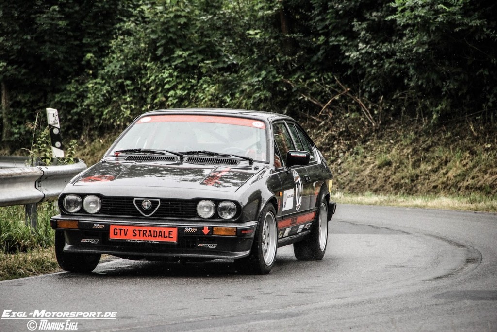 Ein klassischer schwarzer Alfa Romeo GTV Stradale mit roten Akzenten nimmt während des Motorsportevents Weserbergpreis 2016 eine scharfe Kurve auf einer nassen, kurvenreichen Waldstraße. Der Hintergrund ist von üppigem Grün geprägt.