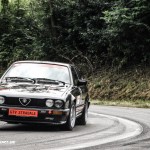 Ein schwarzer Alfa Romeo GTV Stradale, der vorne ein rotes Nummernschild mit dem Modellnamen trägt, fährt auf einer kurvigen Waldstraße. Während das Auto anmutig durch die Kurven navigiert, werden Erinnerungen an seine Leistung beim Weserbergpreis 2016 wach.