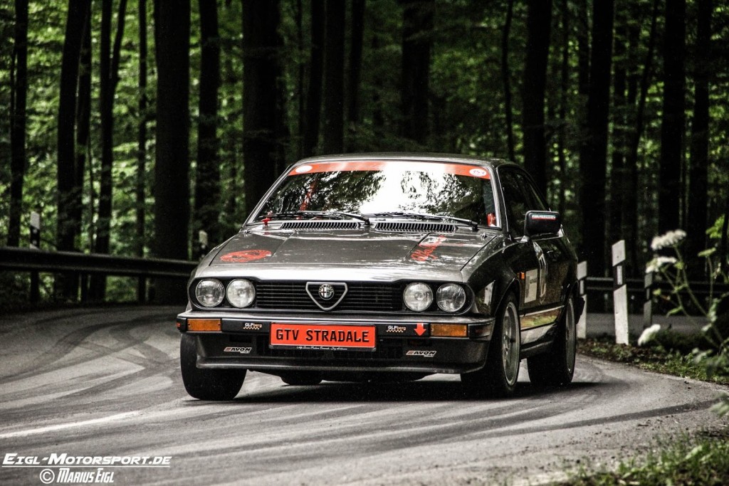 Ein klassischer Alfa Romeo GTV Stradale mit markantem Nummernschild an der Vorderseite navigiert beim Weserbergpreis 2016 über eine kurvenreiche Waldstraße. Die Szene ist dynamisch und spiegelt die Bewegung des Autos und den dichten Wald im Hintergrund wider.