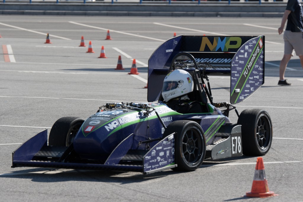 Eine Person mit Helm fährt einen Formula Student-Rennwagen mit der Aufschrift E18 und Sponsorenlogos auf einer mit orangefarbenen Pylonen markierten Rennstrecke.