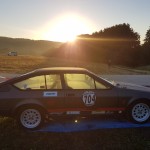 Ein Oldtimer-Rennwagen mit der Nummer 704 auf der Seite parkt bei Sonnenuntergang auf einer blauen Plane neben einer Landstraße.