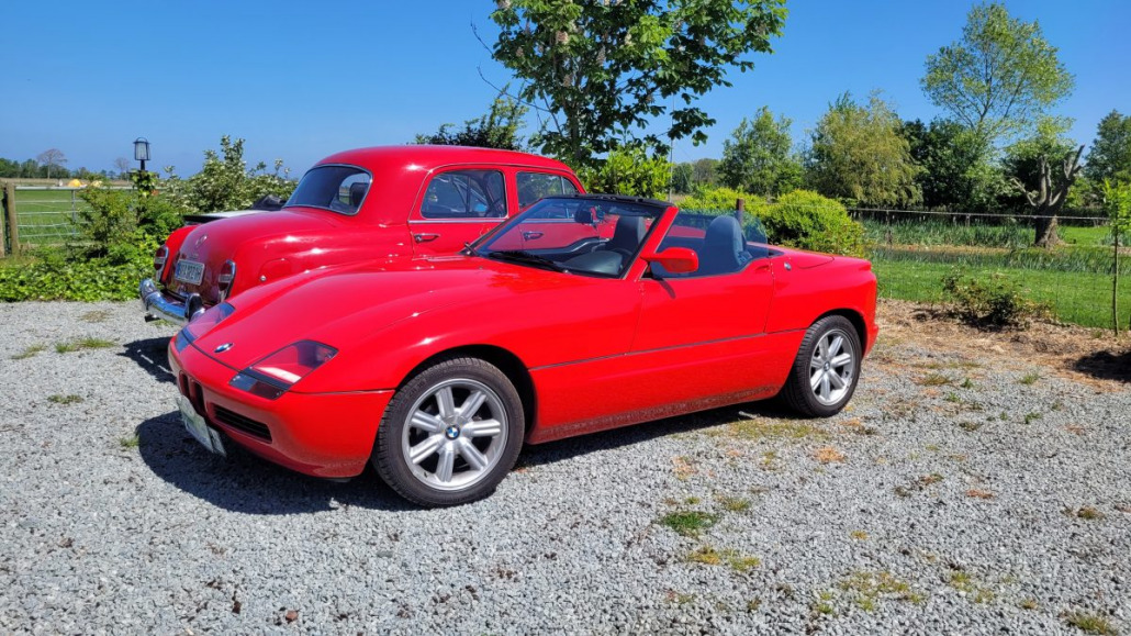 Ein rotes Sportcabriolet steht vor einem roten Oldtimer auf einer Kiesauffahrt, im Hintergrund sind Grünflächen und Bäume zu sehen.