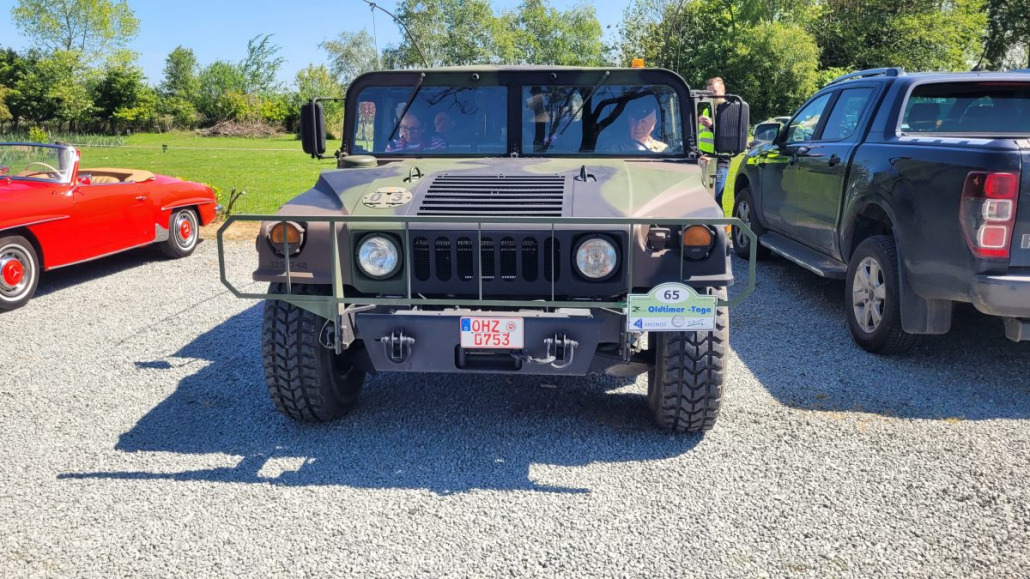 Ein Humvee in Tarnfarbe steht auf Schotter zwischen einem roten Cabrio und einem dunkelfarbigen Pickup vor einer Außenkulisse mit grünen Bäumen im Hintergrund.