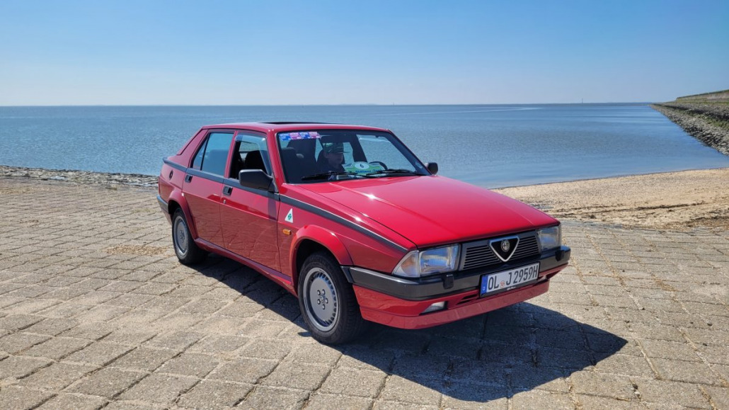 Ein roter Oldtimer steht auf einer gepflasterten Fläche in der Nähe eines Gewässers, im Hintergrund ist ein klarer blauer Himmel zu sehen.