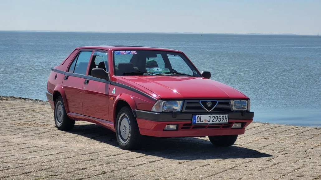 Eine rote Alfa Romeo Limousine parkt auf einer gepflasterten Fläche am Wasser, im Hintergrund ist ein klarer Himmel zu sehen.