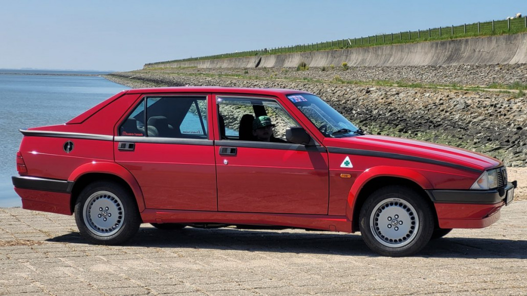 Roter Oldtimer, der auf einer gepflasterten Fläche neben einem Gewässer geparkt ist, mit einem Steindamm und einer Grasfläche im Hintergrund.