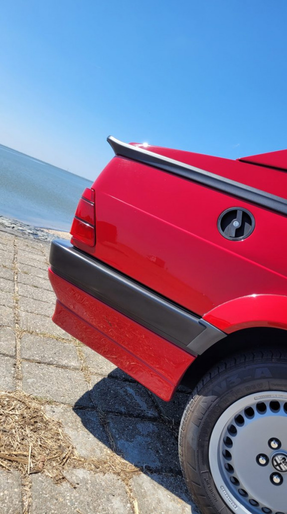 Nahaufnahme der Rückseite eines roten Autos mit Spoiler, das auf einem Steinpflaster neben einem Gewässer unter einem klaren blauen Himmel geparkt ist. Der Fokus liegt auf dem Rad und dem Tankdeckel des Autos.