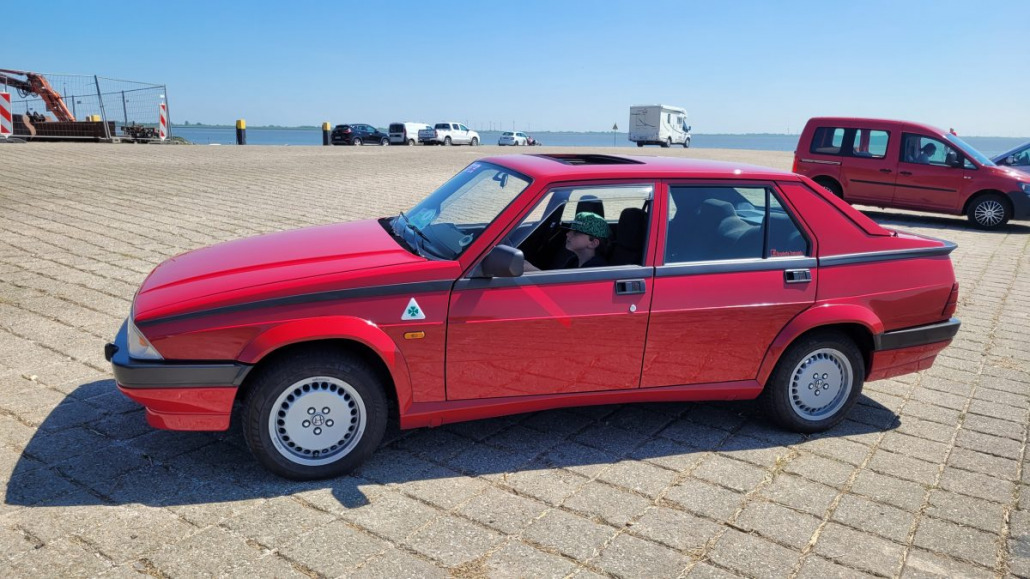 Eine rote Limousine steht auf einer gepflasterten Fläche in der Nähe eines Gewässers. Der Fahrer sitzt im Wageninneren, die Tür ist geschlossen. Im Hintergrund sind mehrere andere Fahrzeuge und eine große Wasserfläche zu sehen.