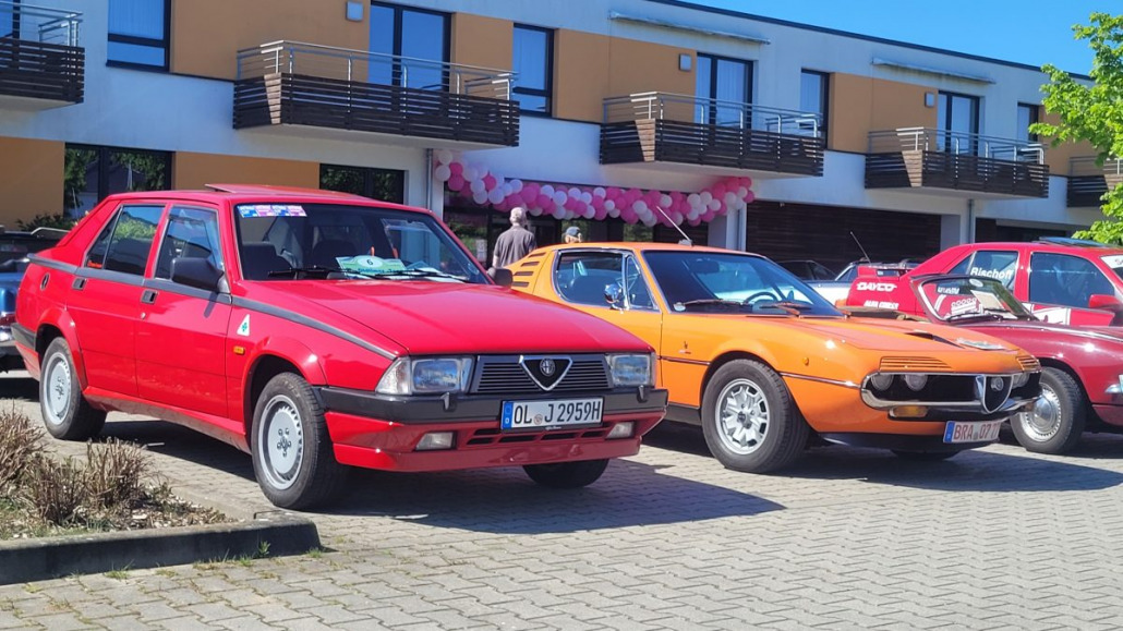 Ein roter und ein orangefarbener Oldtimer stehen nebeneinander auf einem Parkplatz vor einem modernen Gebäude mit Balkonen und rosa Verzierungen.