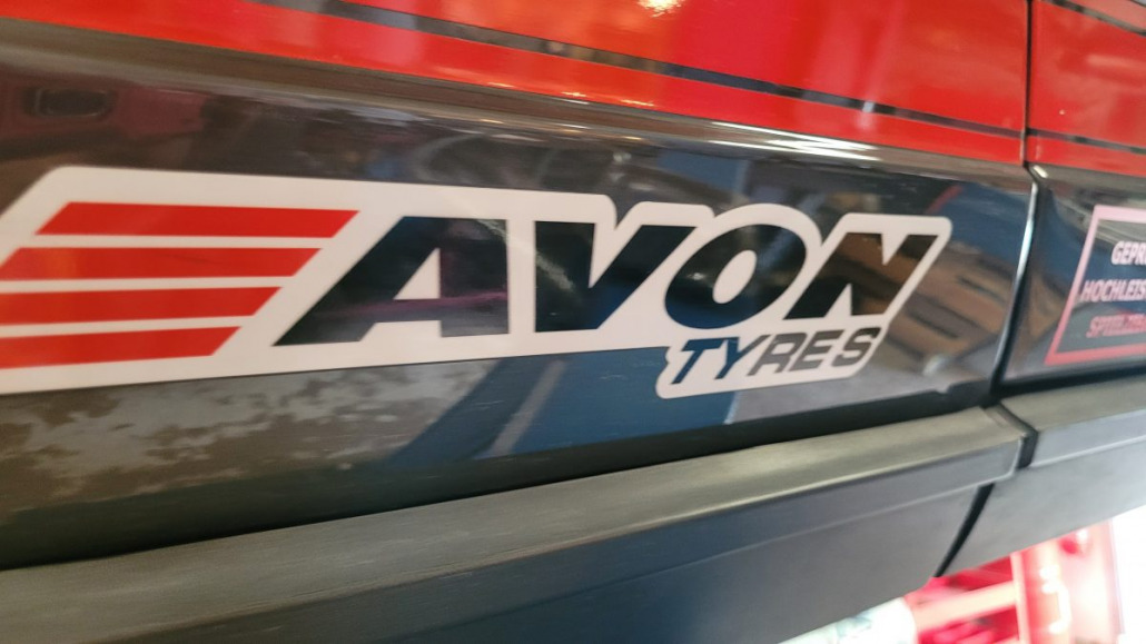 Nahaufnahme eines „Avon Tyres“-Logos an der Seite eines Fahrzeugs. Der Markenname ist in fetten Buchstaben mit roten und weißen Akzenten dargestellt.