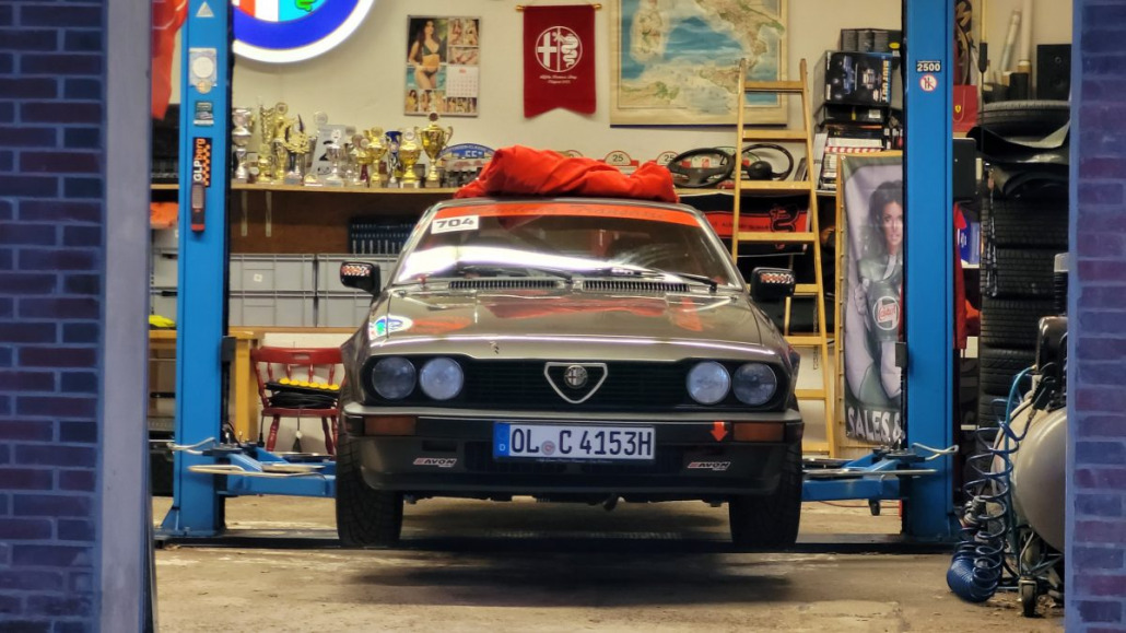 Ein Oldtimer mit dem Nummernschild „OL-C 4153H“ steht auf einer Hebebühne in einer Garage voller Trophäen, Werkzeuge und Erinnerungsstücke aus der Automobilbranche.