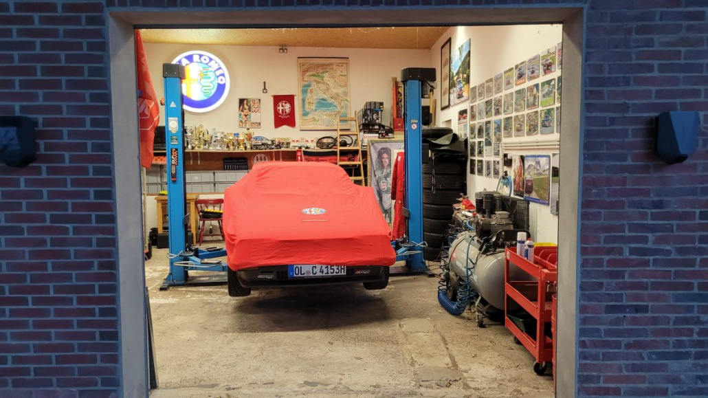 Ein mit einem roten Tuch bedecktes Auto steht in einer Garage voller Werkzeuge, Autoreifen und Motorsport-Erinnerungsstücke. Es gibt zwei Fahrzeughebebühnen und verschiedene Bilder schmücken die Wände.