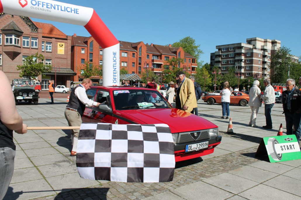 Ein rotes Auto ist gerade dabei, ein Rennen zu starten, während eine Person eine karierte Flagge schwenkt. Im Hintergrund sind auf einem Stadtplatz weitere Autos und Zuschauer zu sehen.