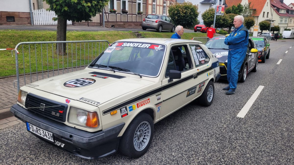 Ein beiges Rennauto mit verschiedenen Aufklebern und der Nummer „700“ auf der Windschutzscheibe steht in einer Reihe auf einer Straße. In der Nähe stehen zwei Männer, einer in einer blauen Jacke und der andere in einem blauen Rennanzug.