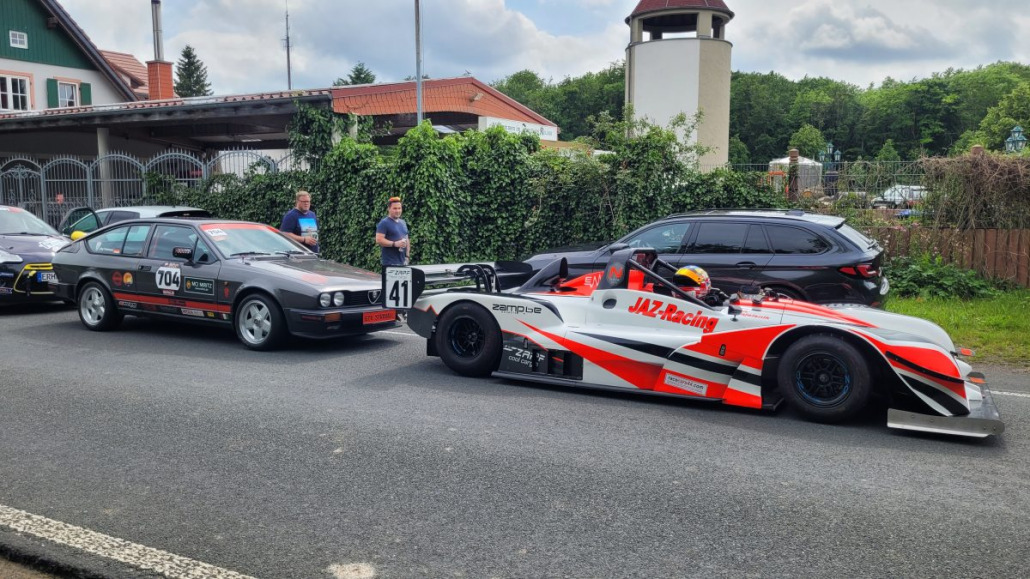 Zwei Rennwagen mit den Nummern 704 und 41 parken auf einer Straße, im Hintergrund sind einige Zuschauer zu sehen. Der Wagen mit der Nummer 41 trägt den Schriftzug „JAZ Racing“ auf der Seite und wirkt dadurch aerodynamischer.