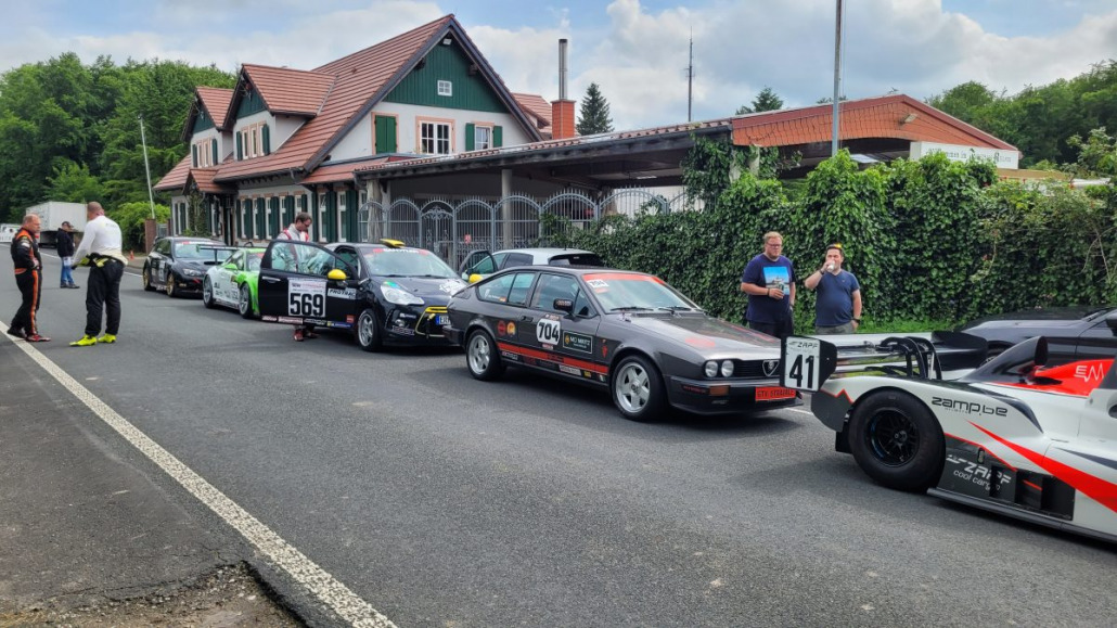 Mehrere Autos stehen auf einer Straße vor einem Haus. Um die Autos herum stehen Männer in Rennkleidung und unterhalten sich. Die Autos tragen unterschiedliche Startnummern wie 569, 704 und 41.