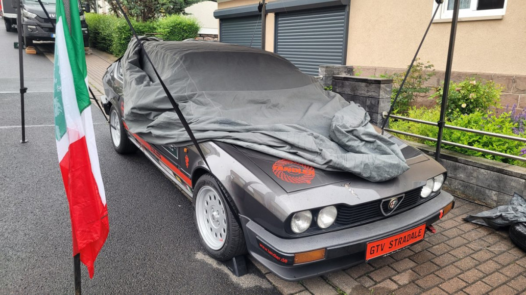 Ein teilweise abgedeckter schwarzer Alfa Romeo GTV Stradale ist auf einer Einfahrt neben einer italienischen Flagge geparkt.