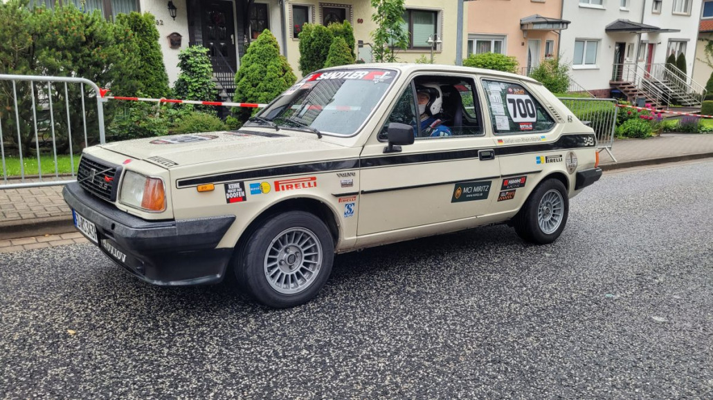 Ein Rallye-Oldtimer mit der Nummer 700 an der Seite steht in einer Wohnstraße und ist mit verschiedenen Rennaufklebern und Sponsoren versehen. Die Straße ist nass, im Hintergrund sind rote Absperrbänder zu sehen.