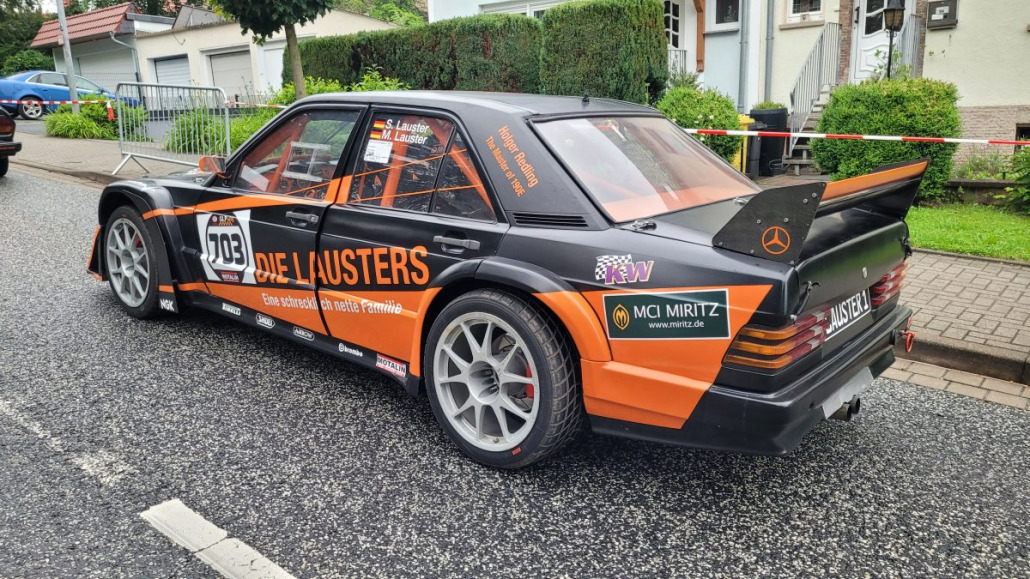 Ein schwarz-orangefarbenes Rennauto mit der Aufklebernummer 103 parkt auf einer Straße. Das Auto ist mit verschiedenen Sponsorenlogos und einem großen Heckspoiler versehen.